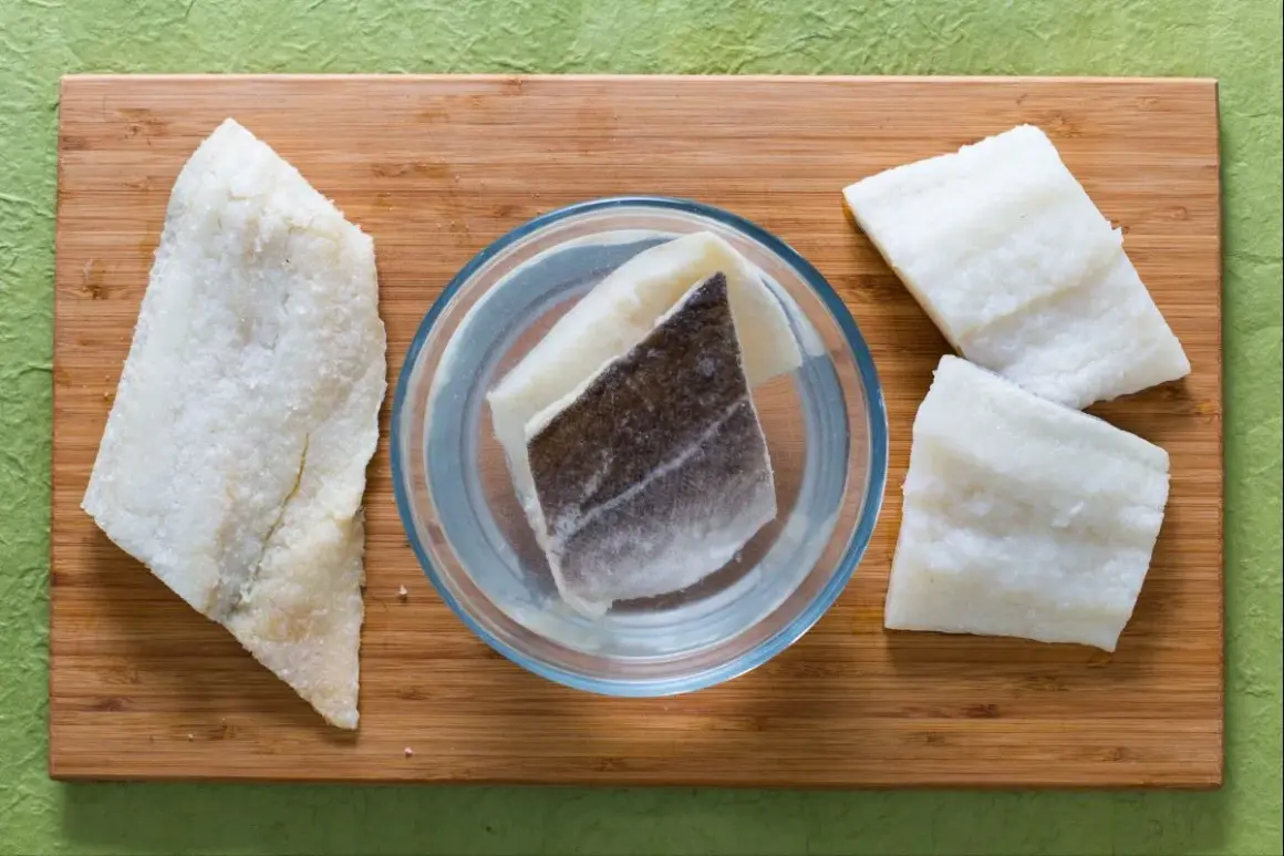 Morue salée prête pour une recette. Des morceaux de poisson sont disposés sur une planche en bois, certains dans un bol d'eau.