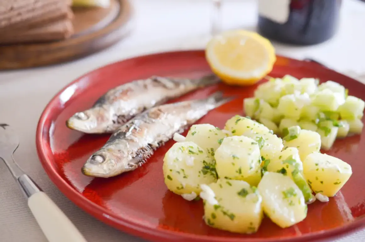 Deux sardines grillées sur une assiette rouge, accompagnées de pommes de terre persillade et d'une salade de concombre. Un vrai délice de sardine barbecue !