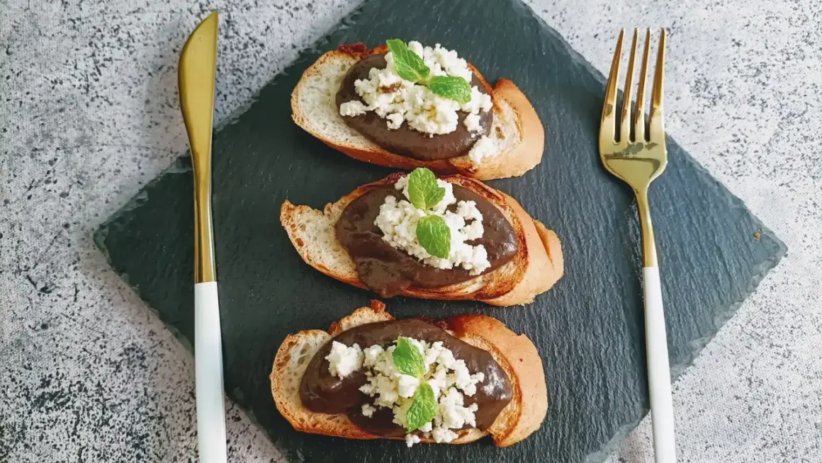 Trois toasts de pain grillé garnis d'une pâte foncée, de fromage blanc émietté et de feuilles de menthe. Une présentation élégante pour une tapenade de la mer revisitée.