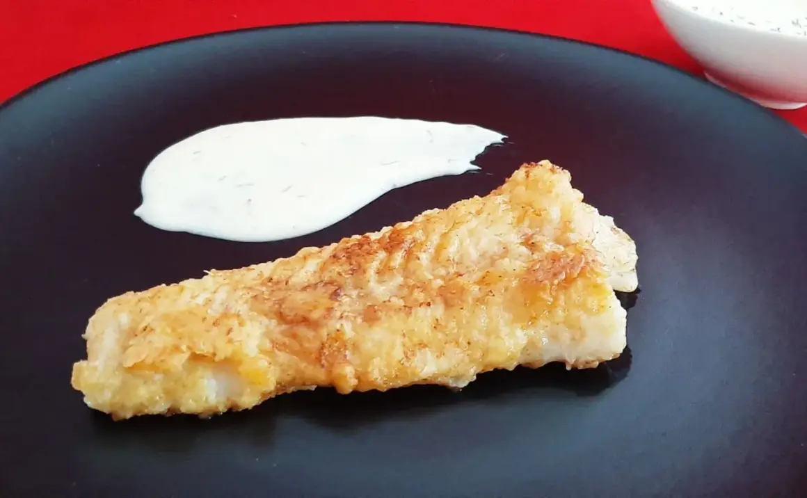 Un morceau de lingue poisson pané, doré et croustillant, servi avec une sauce blanche crémeuse sur une assiette noire.