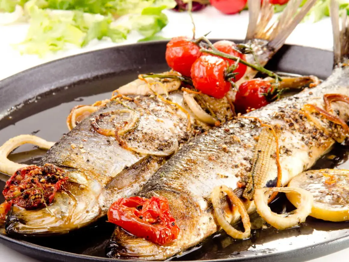 Deux poissons entiers, le hareng, rôtis avec des tomates cerises, des oignons et des rondelles de citron sur un plat noir.