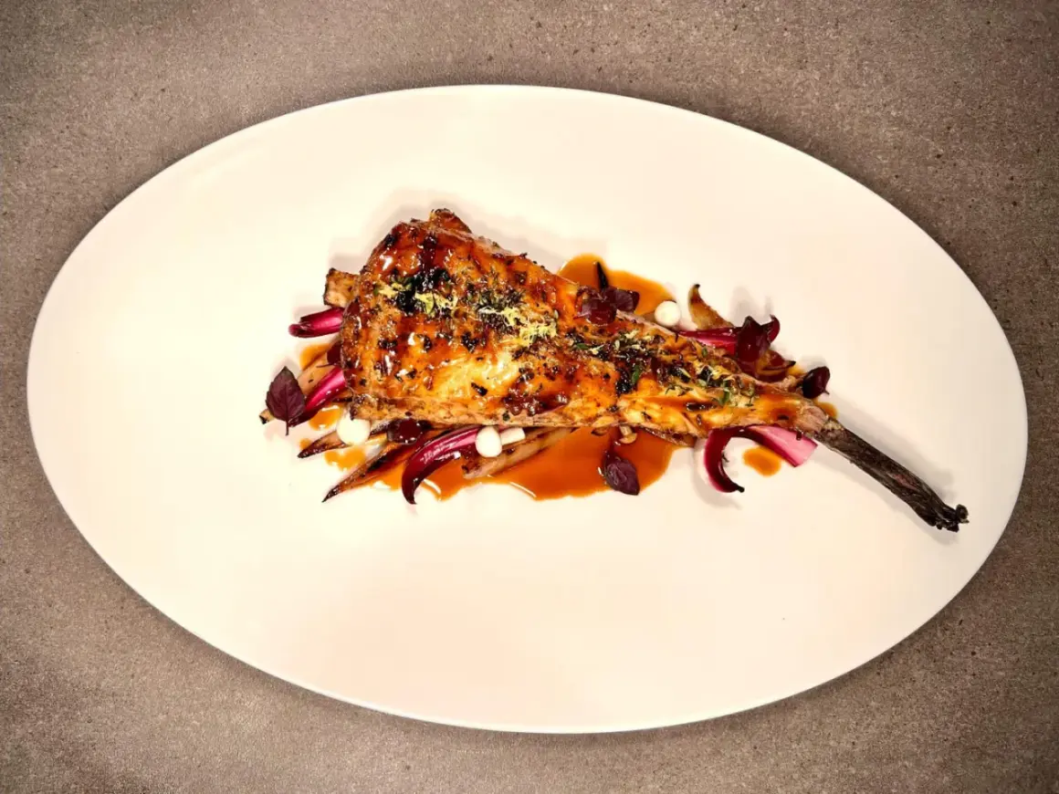 Un plat de lotte au barbecue, laqué et garni d'herbes, servi sur un lit de légumes rouges et blancs.