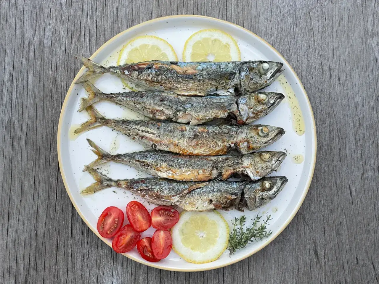Cinq chinchards poissons grill&eacute;s, servis sur une assiette avec des quartiers de citron, des tomates cerises et des herbes.