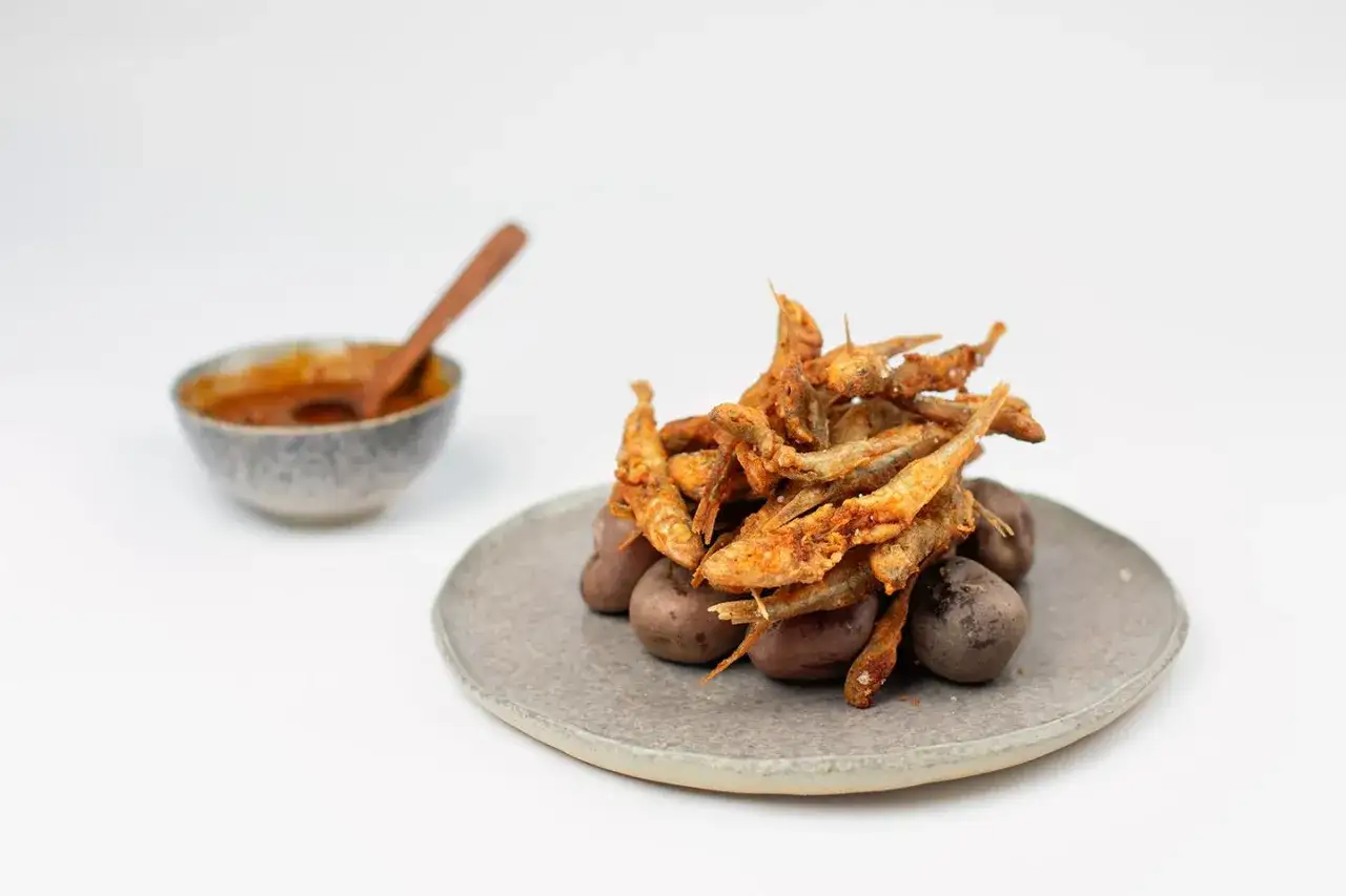 Un tas de petits poissons croustillants, l'&eacute;perlan frit, repose sur des pommes de terre violettes. Un bol de sauce &eacute;pic&eacute;e est &agrave; gauche.