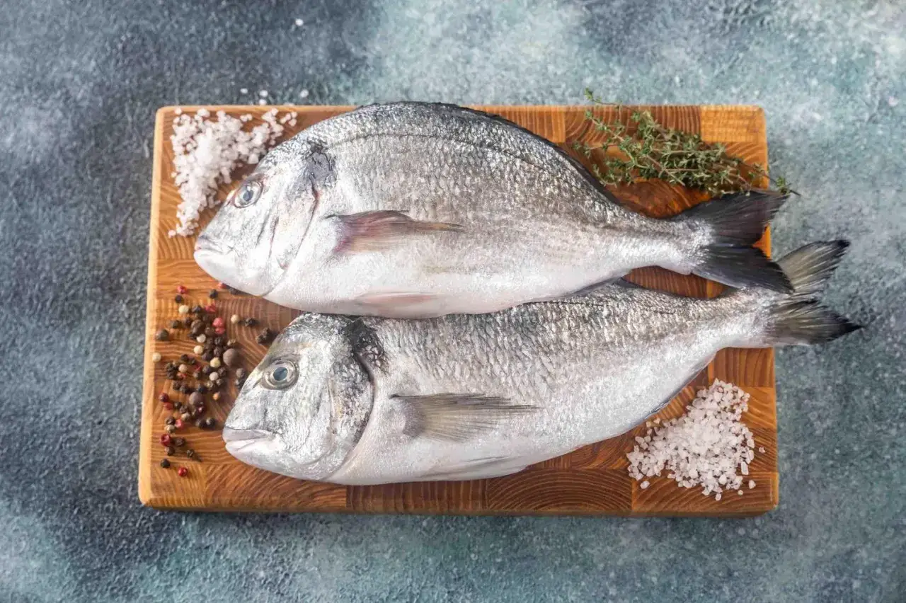 Deux dorades grises entières, prêtes à être cuisinées, sur une planche en bois avec du gros sel, des grains de poivre et une branche de thym.