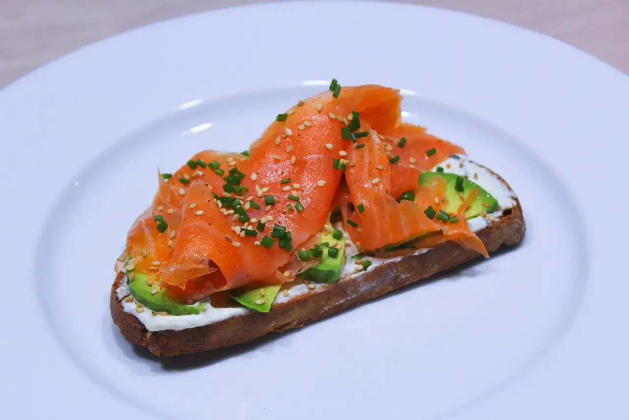 Un toast avocat saumon, garni de graines de s&eacute;same et de ciboulette, sur une assiette blanche.