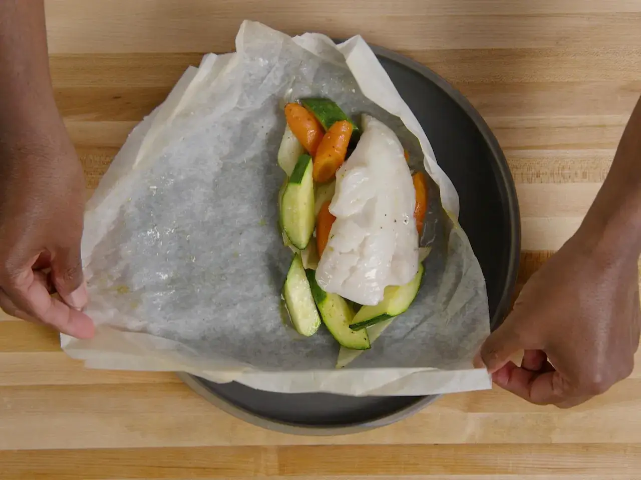 Pr&eacute;paration d'un poisson en papillote avec des l&eacute;gumes frais. Les mains replient d&eacute;licatement le papier sulfuris&eacute;.