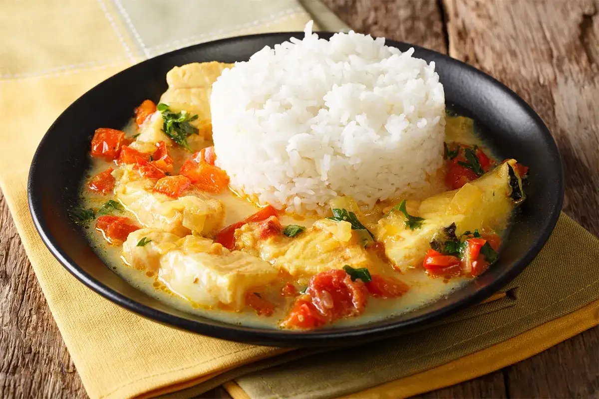 Dos de cabillaud en sauce cr&eacute;meuse avec des tomates et du riz blanc, servi dans une assiette noire sur une serviette jaune.