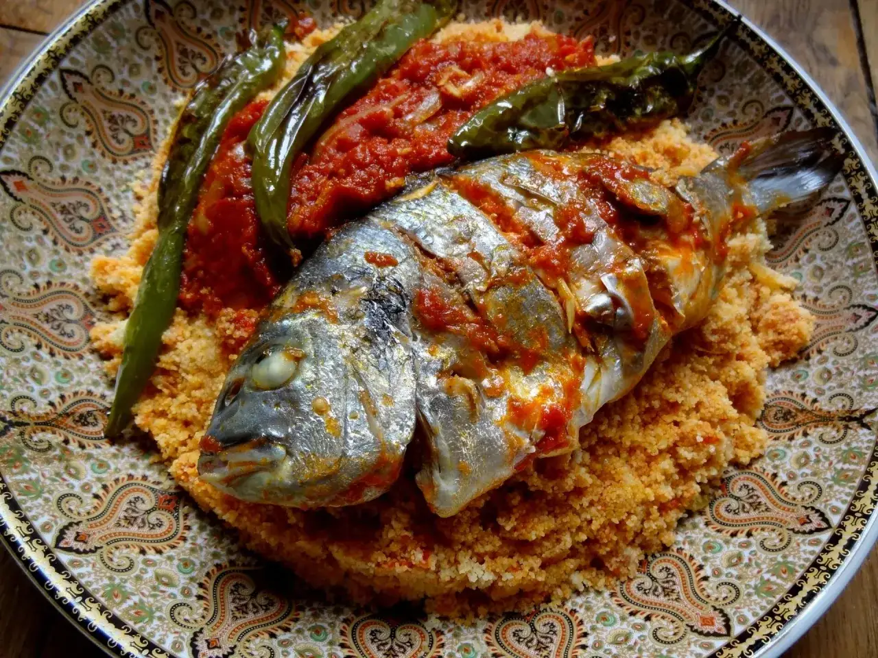 Un plat de couscous poisson, avec un poisson entier nappé de sauce tomate, accompagné de poivrons verts grillés, le tout servi sur une assiette décorée.