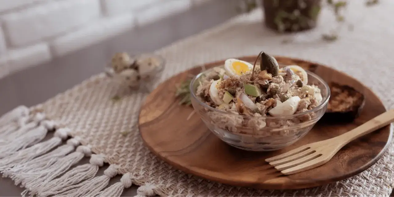 D&eacute;licieuse salade de thon garnie d'&oelig;ufs durs, de c&acirc;pres et d'avocat, servie dans un bol en verre sur une assiette en bois.