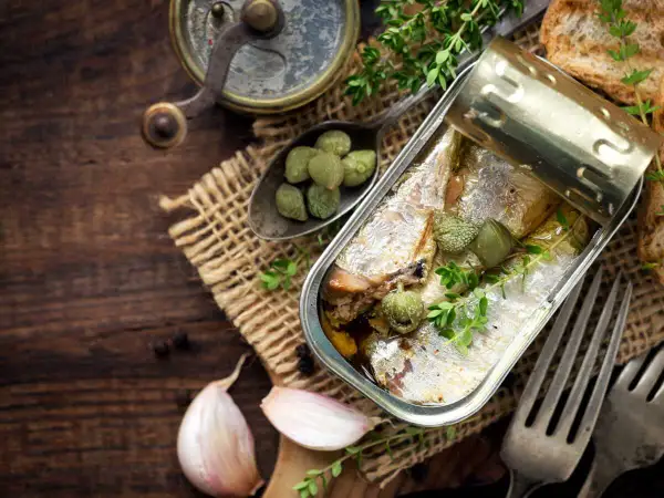 Une boîte ouverte de la sardine, garnie de câpres et de thym, sur un fond de bois rustique avec de l'ail et des toasts.