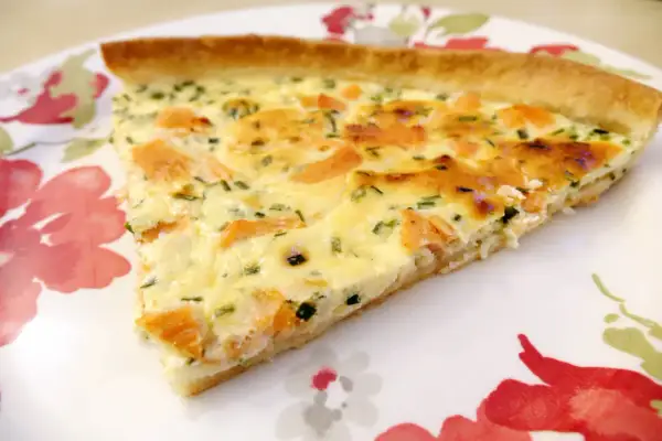 Une part de quiche au saumon fumé, garnie d'herbes fraîches, sur une assiette décorée de fleurs rouges et vertes.