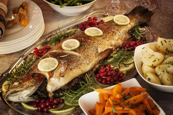 Un poisson entier, un turbot, est servi sur un plateau avec des rondelles de citron, des airelles et du romarin. Des pommes de terre et des carottes sont disposées sur les côtés.