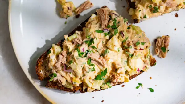 Une tranche de pain noir garnie d'une généreuse salade de thon crémeuse, avec des morceaux d'œuf dur, de céleri et d'oignon rouge, parsemée de persil frais.