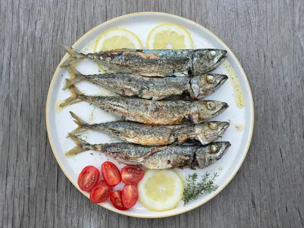 Cinq chinchards poissons grillés, servis sur une assiette avec des quartiers de citron, des tomates cerises et des herbes.