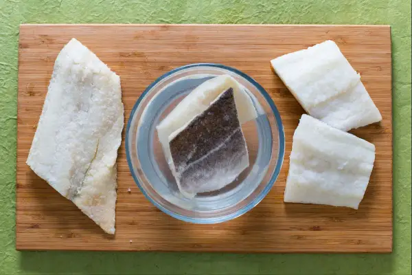 Morue salée prête pour une recette. Des morceaux de poisson sont disposés sur une planche en bois, certains dans un bol d'eau.