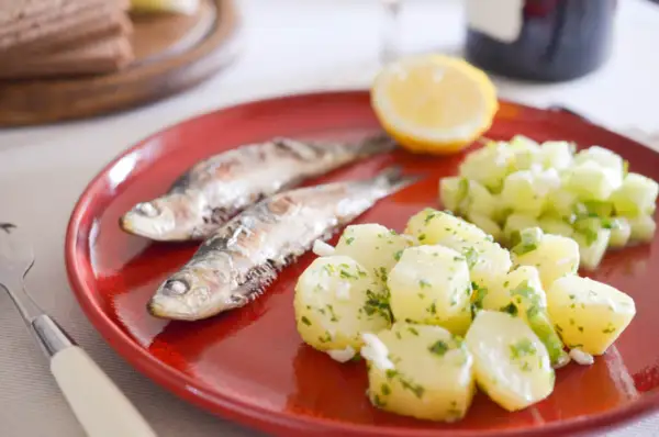 Deux sardines grillées sur une assiette rouge, accompagnées de pommes de terre persillade et d'une salade de concombre. Un vrai délice de sardine barbecue !