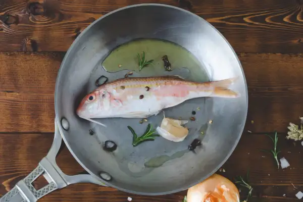 Un poisson rouge prêt à être cuisiné à la poêle avec de l'ail, du romarin et des grains de poivre sur une planche en bois.