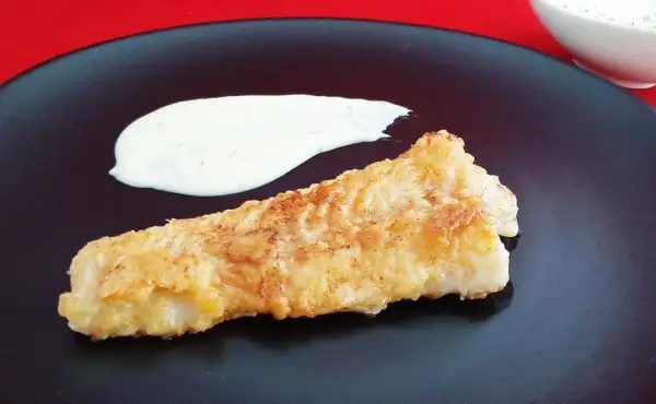 Un morceau de lingue poisson pané, doré et croustillant, servi avec une sauce blanche crémeuse sur une assiette noire.