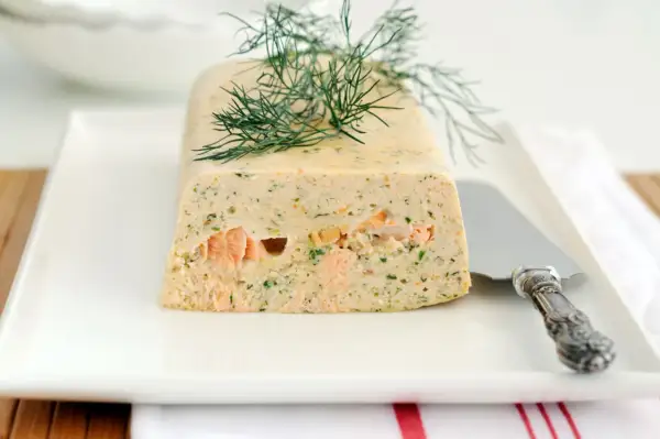 Terrine de saumon frais et fumé garnie d'aneth sur une assiette blanche avec un couteau.