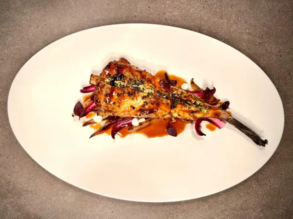 Un plat de lotte au barbecue, laqué et garni d'herbes, servi sur un lit de légumes rouges et blancs.