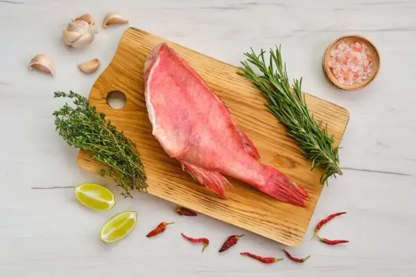 Un sébaste poisson cru, prêt à être cuisiné avec ail, thym, romarin, piments et sel rose sur une planche en bois.