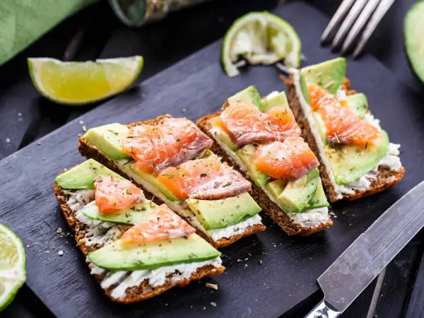 Quatre toasts avocat saumon, garniture crémeuse, herbes et quartiers de citron vert sur planche noire.
