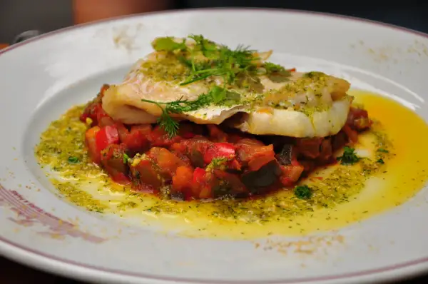 Un délicieux dos de cabillaud servi sur un lit de légumes méditerranéens, arrosé d'une sauce verte et d'huile d'olive.